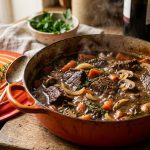 Rich beef casserole in an orange Le Creuset cast iron pot with carrots, mushrooms and herbs in red wine gravy