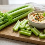 Fresh celery stalks on a wooden board with sliced pieces and a bowl of hummus for dipping