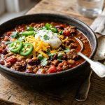 Rich beef chili con carne in a dark ceramic bowl topped with sour cream, cheddar, jalapeños and coriander