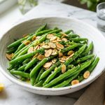 Crisp cooked green beans in a white bowl with toasted almonds and fresh lemon on a marble surface