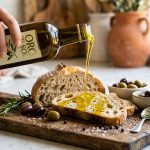 Bottle of extra virgin olive oil being drizzled over rustic bread and olives on a wooden board