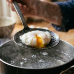 Perfectly poached egg on a slotted spoon over steaming water with runny yolk visible